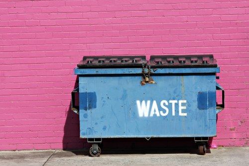 A large blue waste skip with a hinged lid, positioned on a concrete surface in front of a vibrant pink brick wall. The skip is made of metal, with visible rust and dirt marks, and has the word 'WASTE' painted in white capital letters on its side. The lid is closed, secured with a metal latch, and wheels are visible at the base for mobility. The surrounding environment appears to be an urban or commercial area, consistent with waste collection and rubbish removal services provided by companies like Commercial Waste Hackney, serving local businesses and premises within the Hackney postcode area. The overall scene emphasizes the functional aspect of waste management infrastructure in a city environment, highlighting the importance of proper rubbish disposal and the role of professional waste removal specialists.