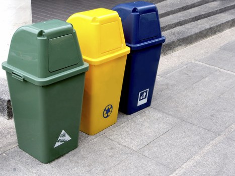 Three upright commercial rubbish bins situated on a paved urban sidewalk, positioned side by side. The first bin on the left is dark green with a slightly textured surface, a rounded lid, and an arrow-shaped recycling symbol on the front. The middle bin is yellow, also with a textured surface and a rounded lid, featuring a circular recycling icon with arrows on the front, suggesting its use for recyclable waste. The third bin on the right is dark blue with a smooth finish, a rounded lid, and a small white label with an icon indicating general rubbish disposal. All three bins are placed on a grey concrete pavement next to a building with dark, ribbed external cladding, and are aligned on the right side of the frame, suggesting a typical waste collection point outside a commercial or residential property in Hackney. The scene is illuminated by natural light, providing clear visibility of the bins' colors, textures, and positioning, reflecting the type of waste management services offered by Commercial Waste Hackney in the area.