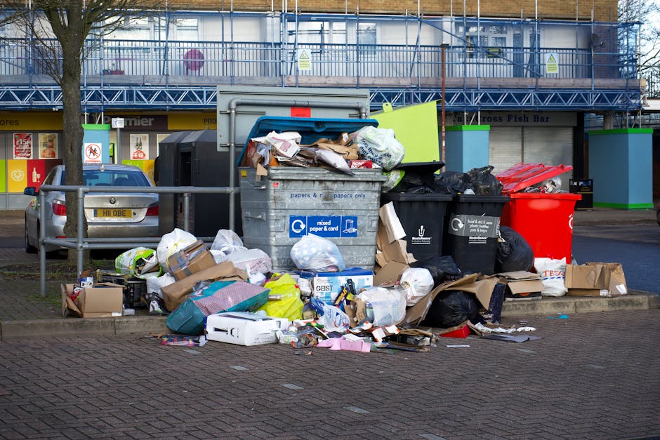A large pile of mixed household waste and recyclables is overflowing from a grey and blue communal rubbish bin situated on a paved urban street. The waste includes crumpled cardboard boxes, plastic ba