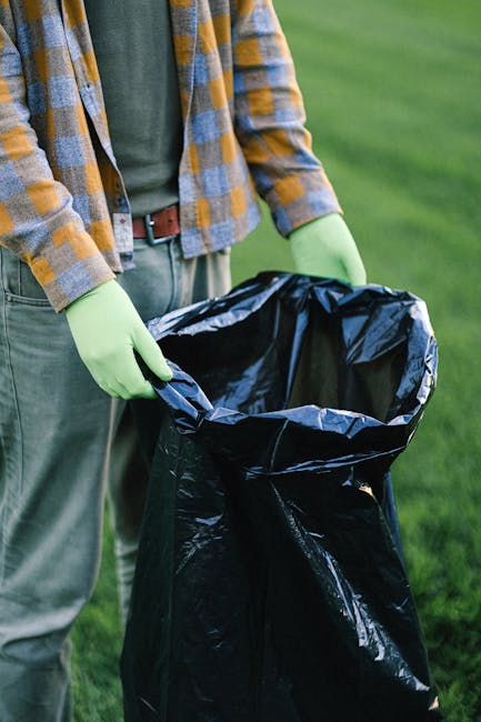 A large pile of mixed household waste and recyclables is overflowing from a grey and blue communal rubbish bin situated on a paved urban street. The waste includes crumpled cardboard boxes, plastic ba
