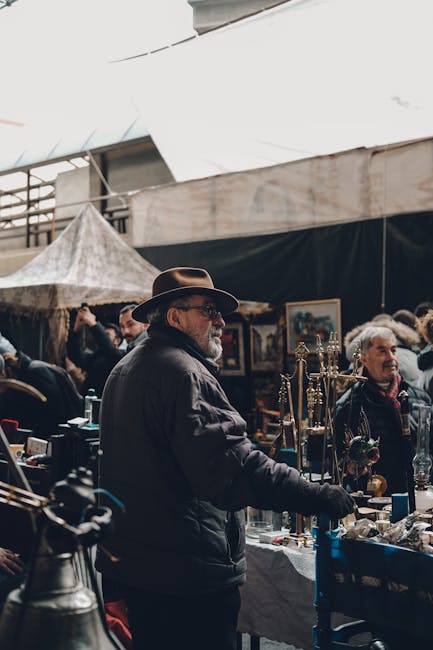 The image depicts a market trader with a grey beard, wearing a wide-brimmed brown hat and dark jacket, standing behind a stall displaying an assortment of antique or vintage items such as candelabras 