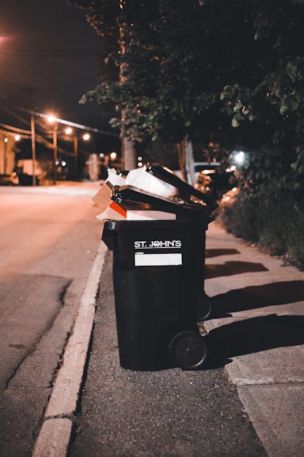 A black wheelie bin with a white label and the name 'St. John's' printed on it stands on a paved sidewalk beside a street at night. The bin is filled with various types of waste, including cardboard b