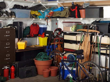 A cluttered garage interior containing various stored items typical of a household storage space near Hackney. In the foreground, several plastic plant pots of different sizes and colors, including terracotta and dark brown, are stacked or placed on the floor. A partially visible blue bicycle with a black seat and handlebars leans against a shelving unit on the right side; the bike's front wheel is turned slightly to the left. To the left of the bicycle, there is a coiled green garden hose resting on the floor. Shelving units above and around hold a variety of objects, such as black plastic storage boxes, a red and black tool bag, yellow and blue buckets, and assorted bags and containers. The top shelves feature fabric or plastic tool rolls, cords, and other miscellaneous items. The environment appears to be a typical garage or storage area used for household rubbish and equipment, with a metal shelving system providing organized storage space, and the overall scene conveying a sense of clutter consistent with rubbish removal and storage management in Hackney or nearby London districts. The lighting suggests natural or ambient source inside a confined space, emphasizing the diversity of stored items.